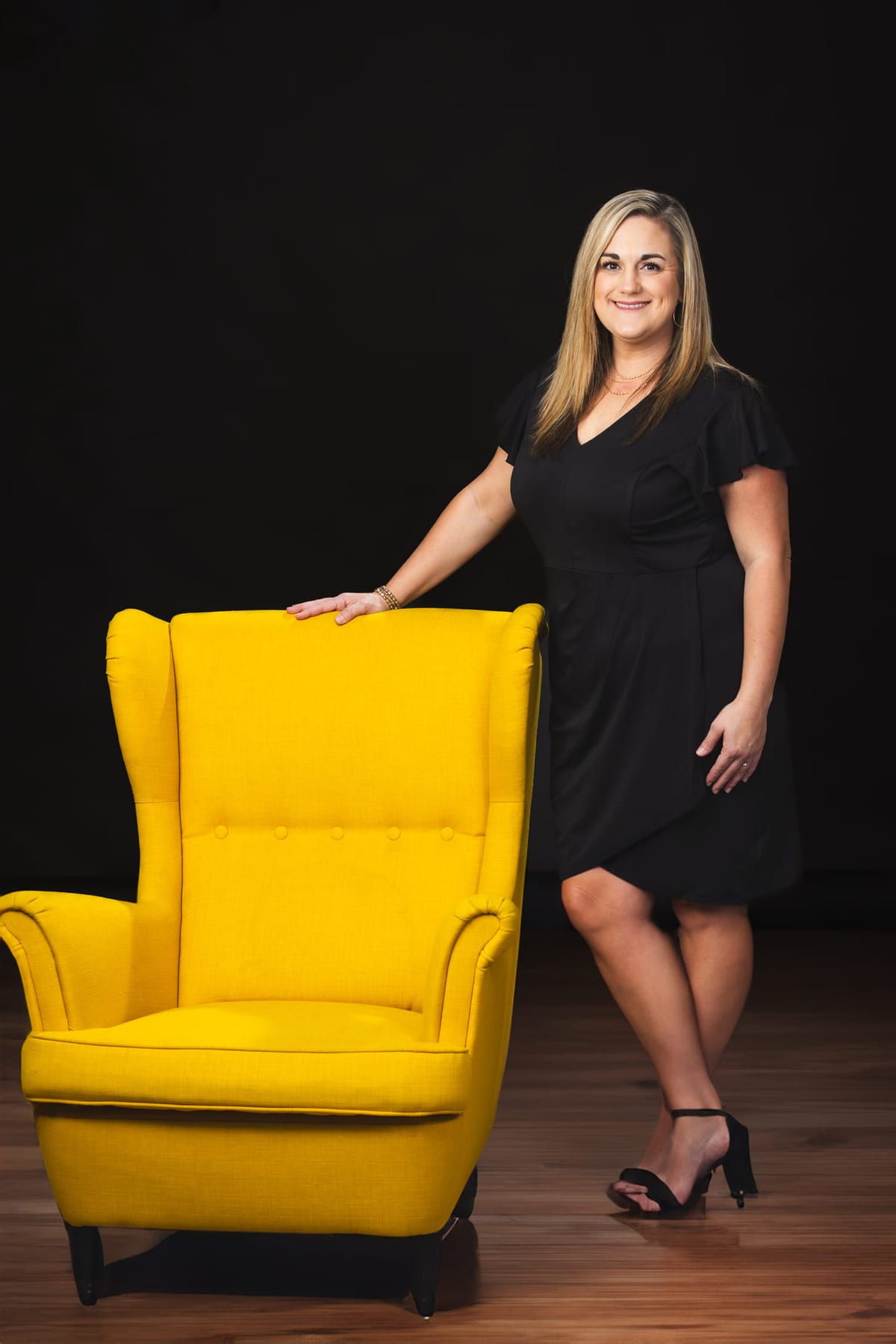 A studio portrait beside a yellow wing chair on a dark set.