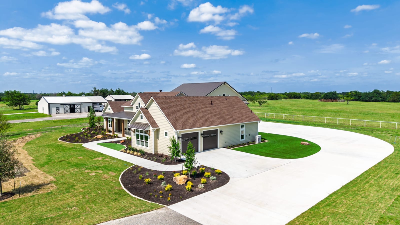 Aerial of a ranch house and outbuilding, a circular drive and flowerbed set in flat green pasture.