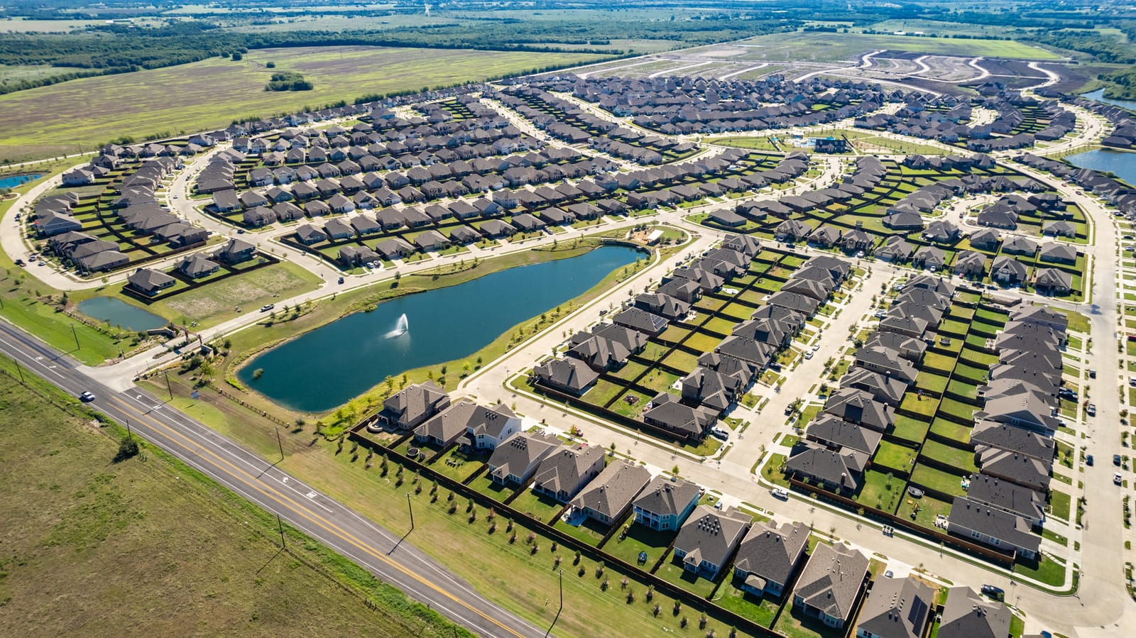 Aerial over a new-build subdivision arranged around a landscaped pond, rooftops running to the horizon.