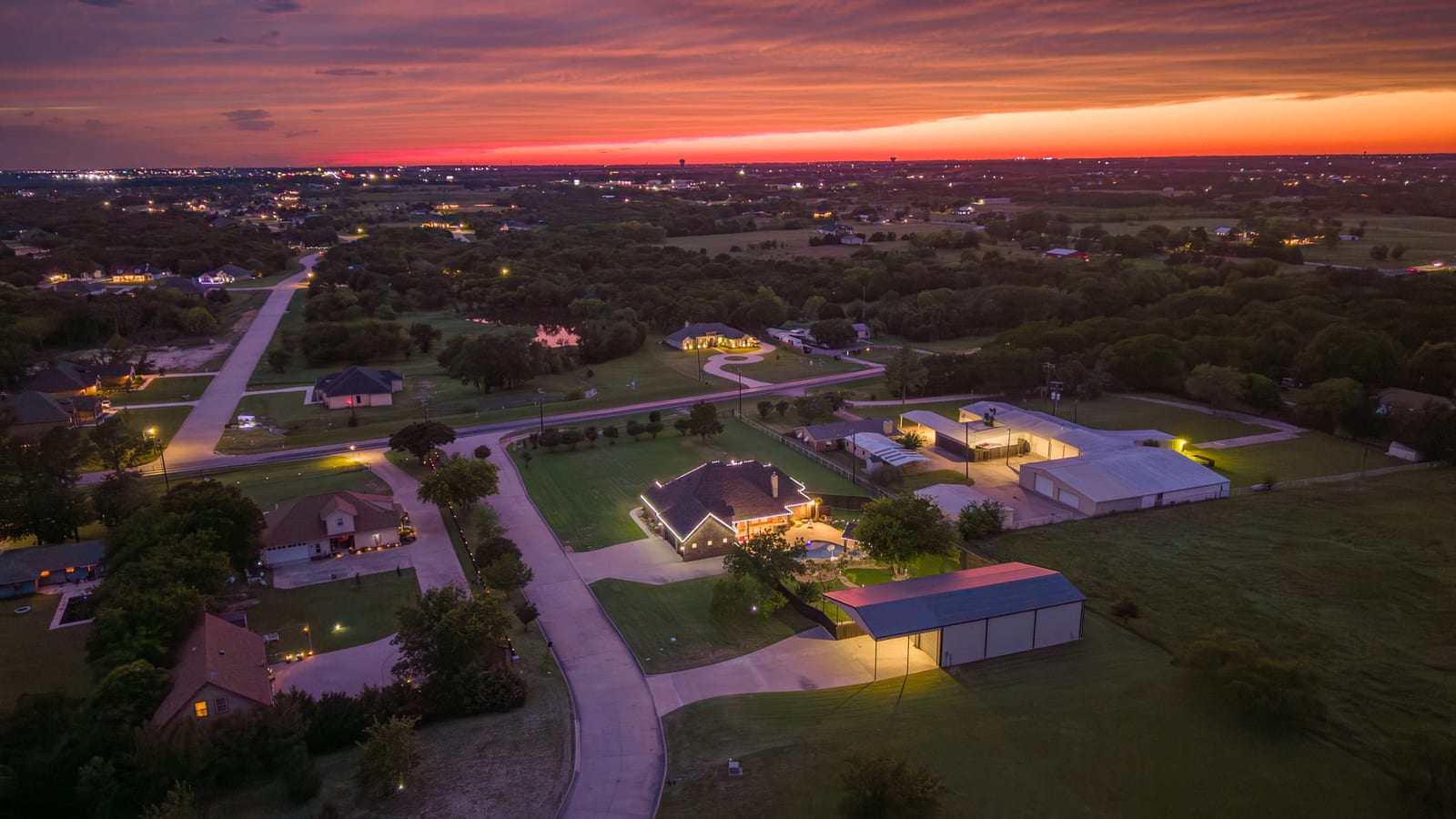 Aerial over rural acreage at sunset, scattered homes and outbuildings under a band of orange sky.
