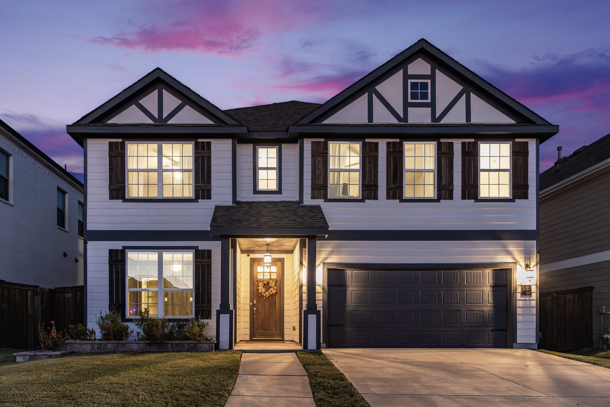 A two-storey house at twilight under a lavender sky, the entry light on over a dark front door and the drive running out of frame.