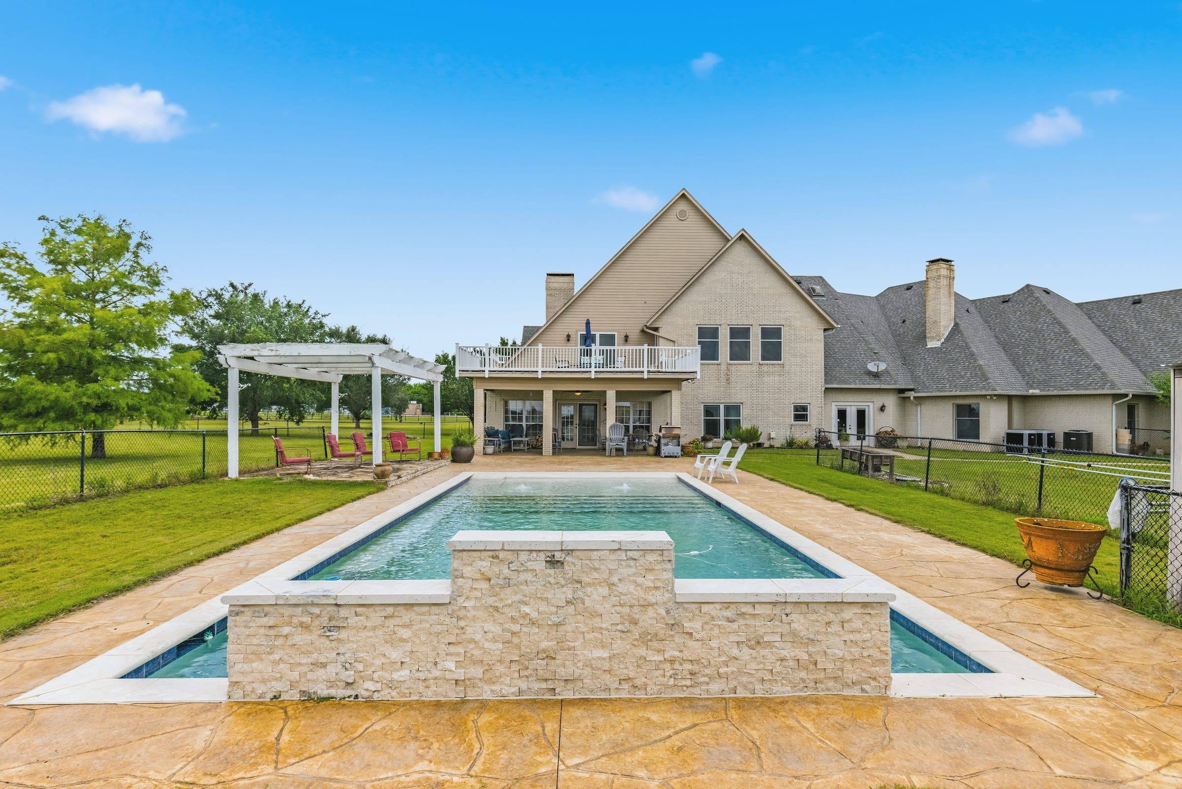 A rectangular pool with a raised stone water feature in the foreground, a pergola and a large farmhouse behind it.