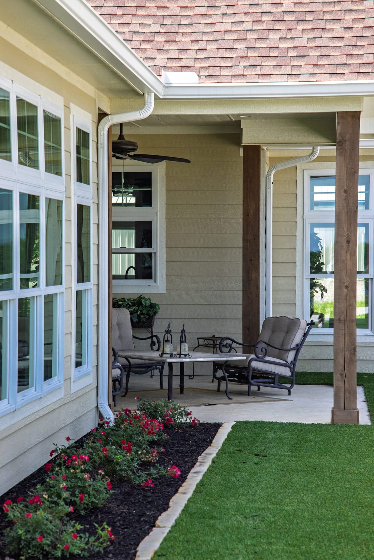 A covered patio with wicker seating and a ceiling fan, roses bedded along the edge of the lawn.