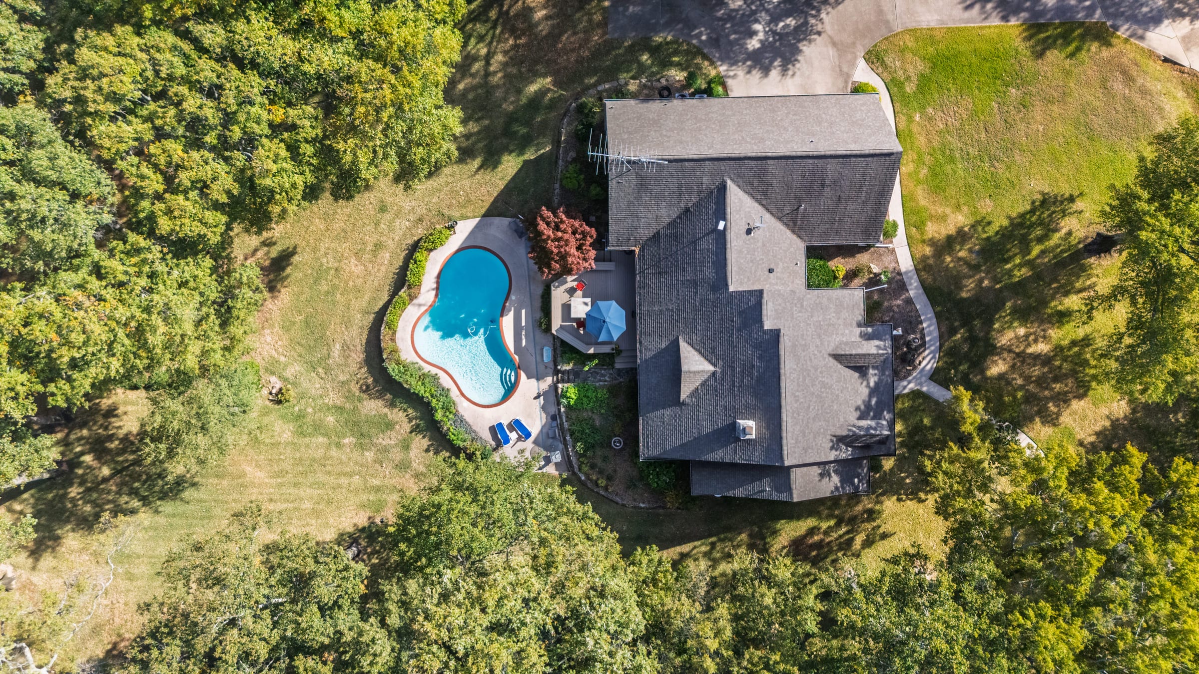 Aerial view looking straight down on a slate-roofed house set in woodland, a kidney-shaped pool and terrace beside it.