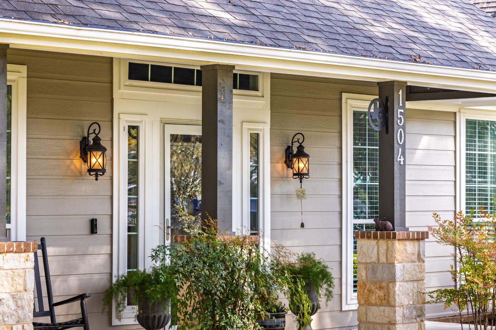 A board-and-batten front porch with rocking chairs, carriage lanterns and potted ferns flanking the door.