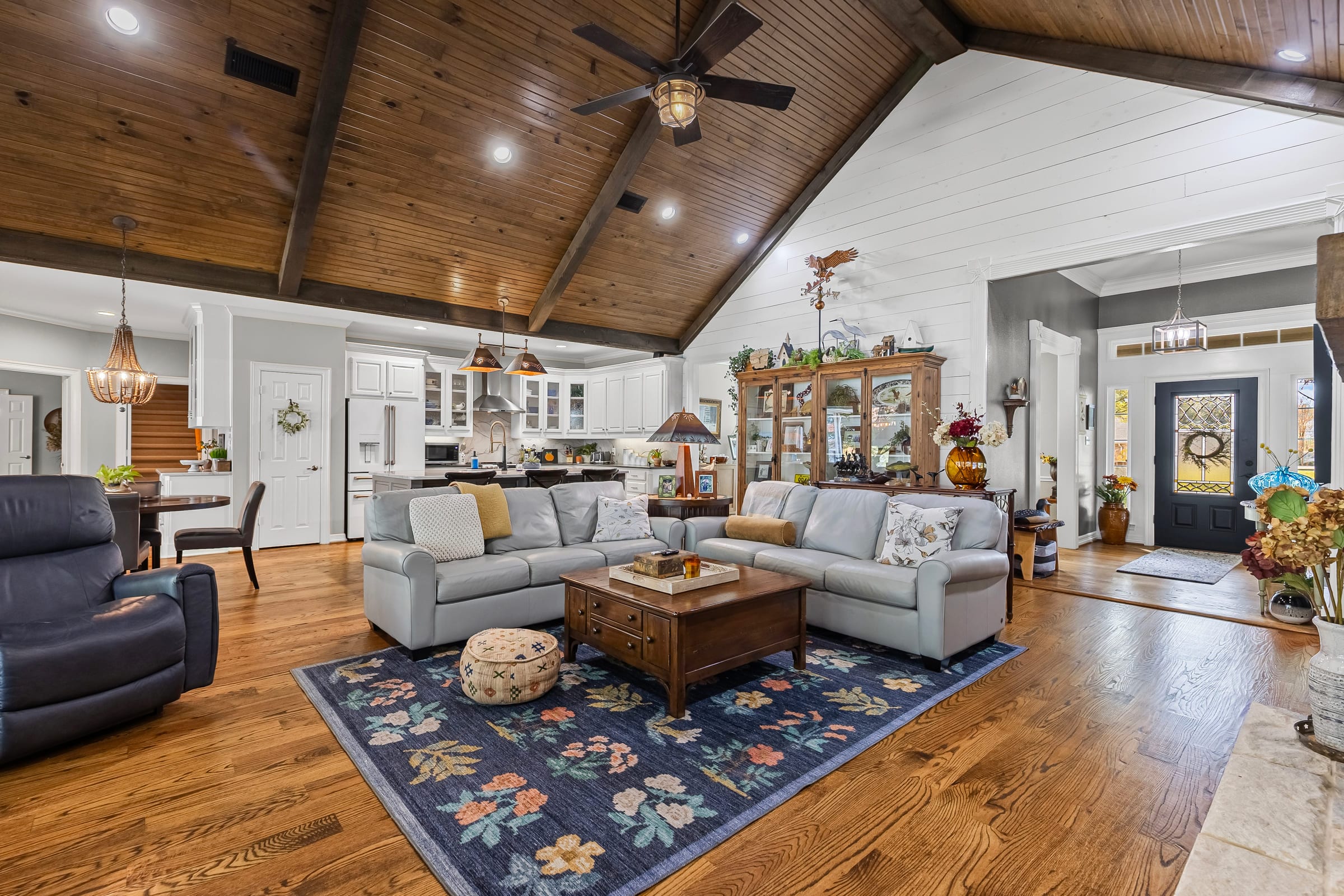 A living room under a vaulted plank ceiling with an exposed beam, seating on a patterned rug and the front door visible past the kitchen.