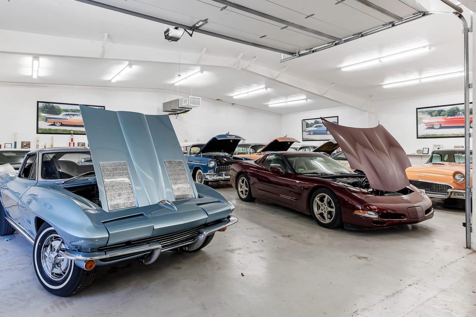 A finished garage with classic Corvettes parked under bright ceiling lights, covers pulled back from the hoods.