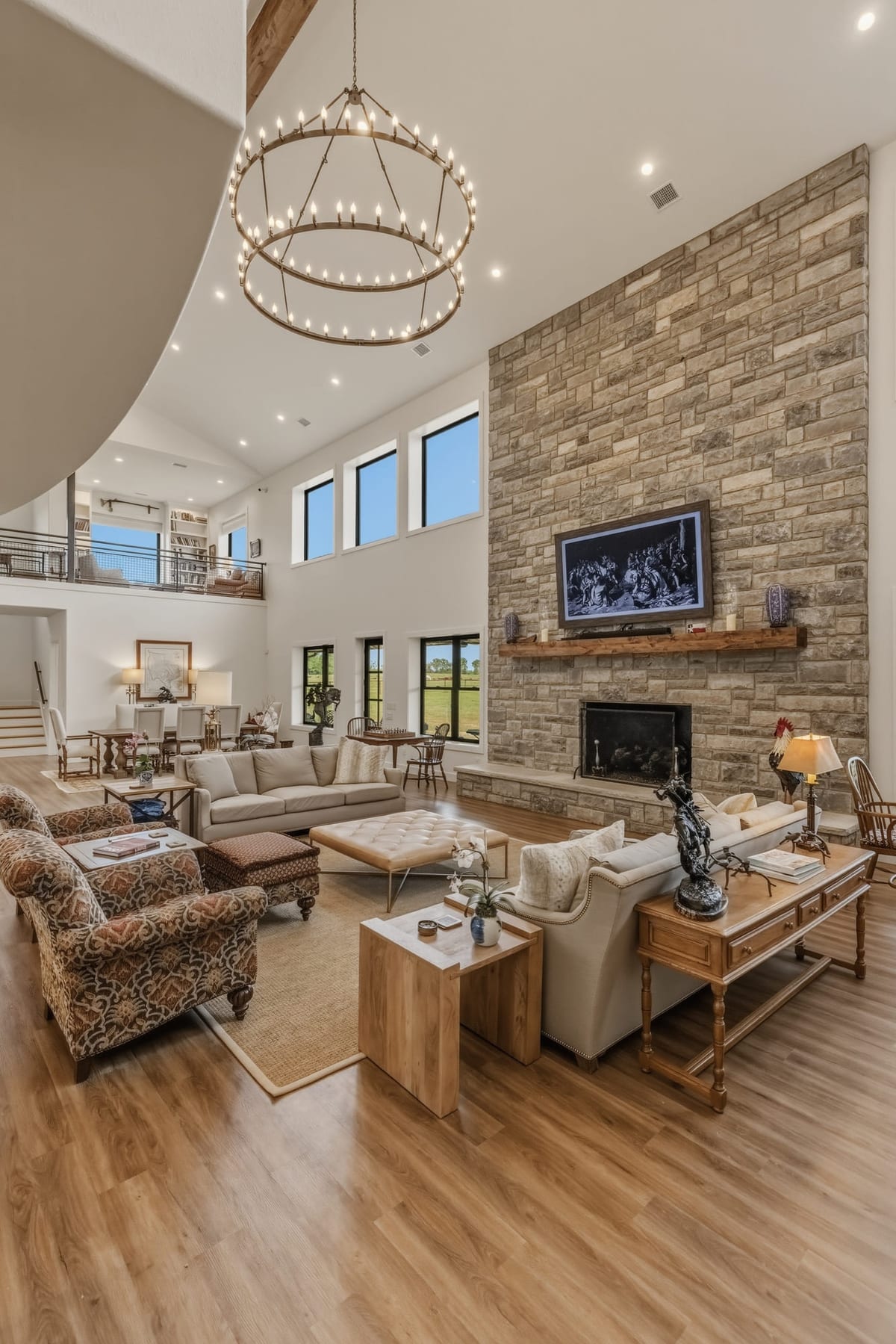 A two-storey great room with a stone chimney breast, iron chandelier and a wall of windows to the field.