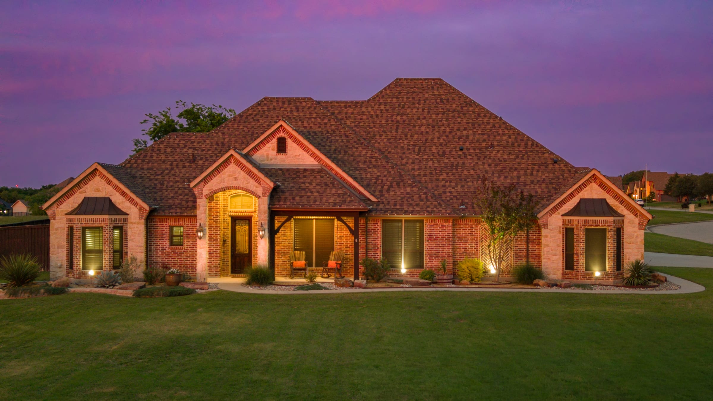 A single-storey brick and stone house photographed at dusk, every window lit warm against a violet sky, lawn running to the edge of the frame.