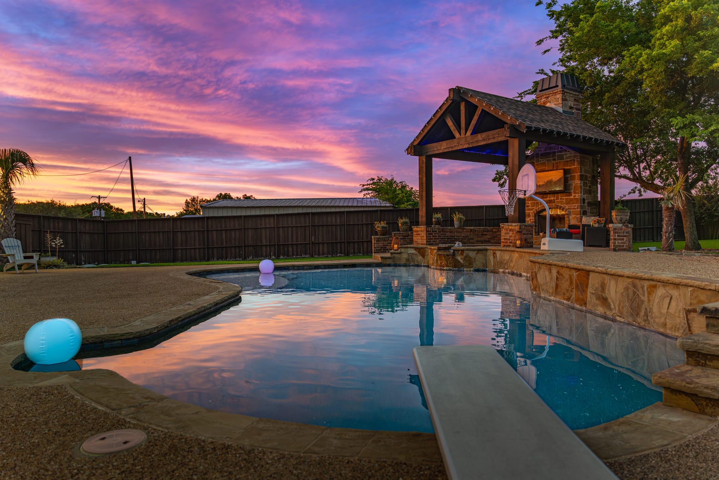 A backyard pool at dusk under a pink and blue sky, a timber-framed outdoor kitchen lit warm at the far end.