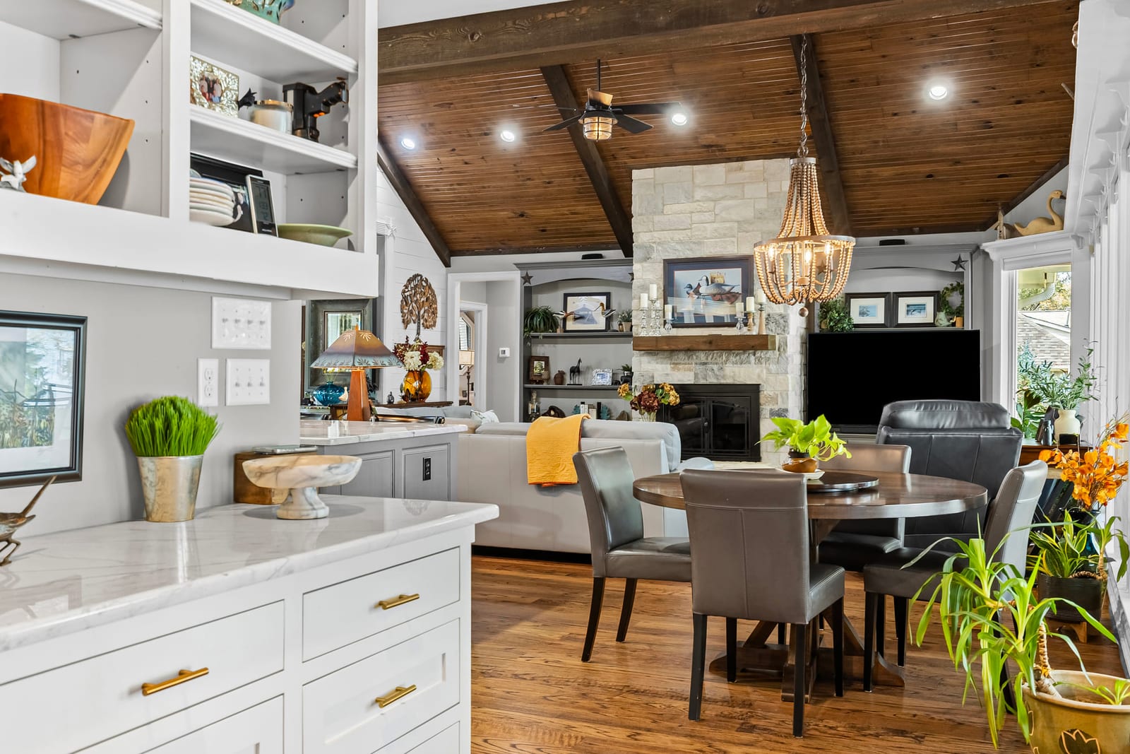 A white kitchen opening onto a breakfast table and living room, a stained plank ceiling running the length of the space.