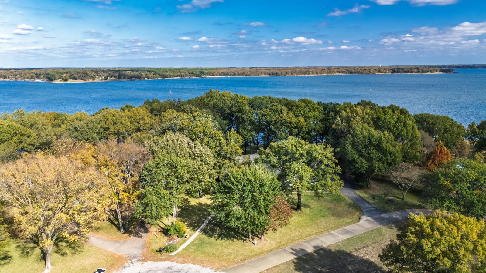 Aerial over wooded lakefront acreage, the open water of the lake filling the horizon behind the treeline.