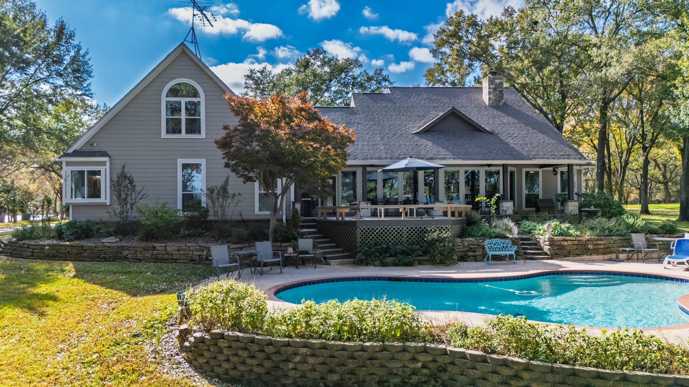 A gabled lake house behind a curving swimming pool and terraced lawn under a bright blue sky.