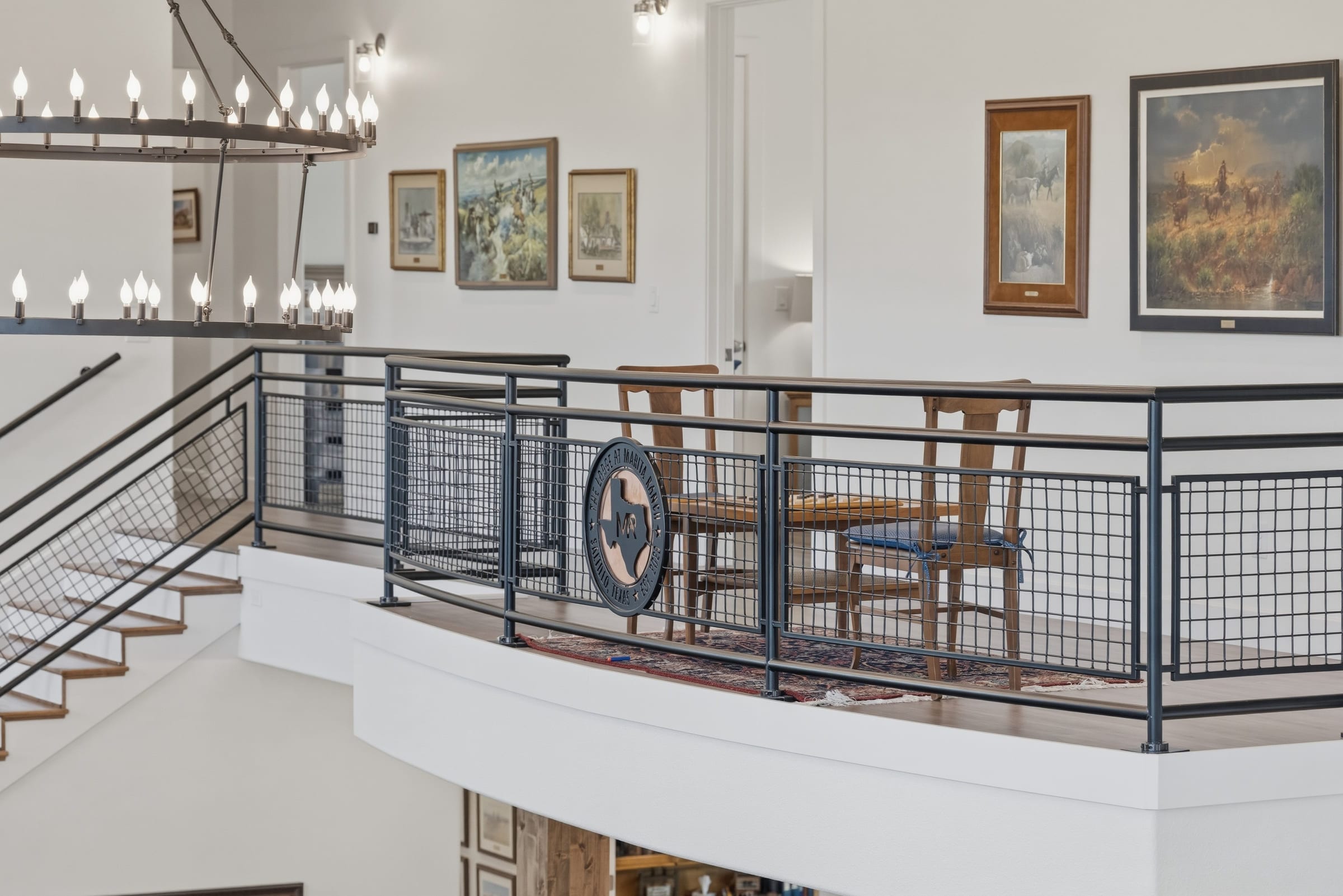 A first-floor landing behind a black wire-and-steel railing, framed pictures hung the length of the wall and an iron chandelier overhead.