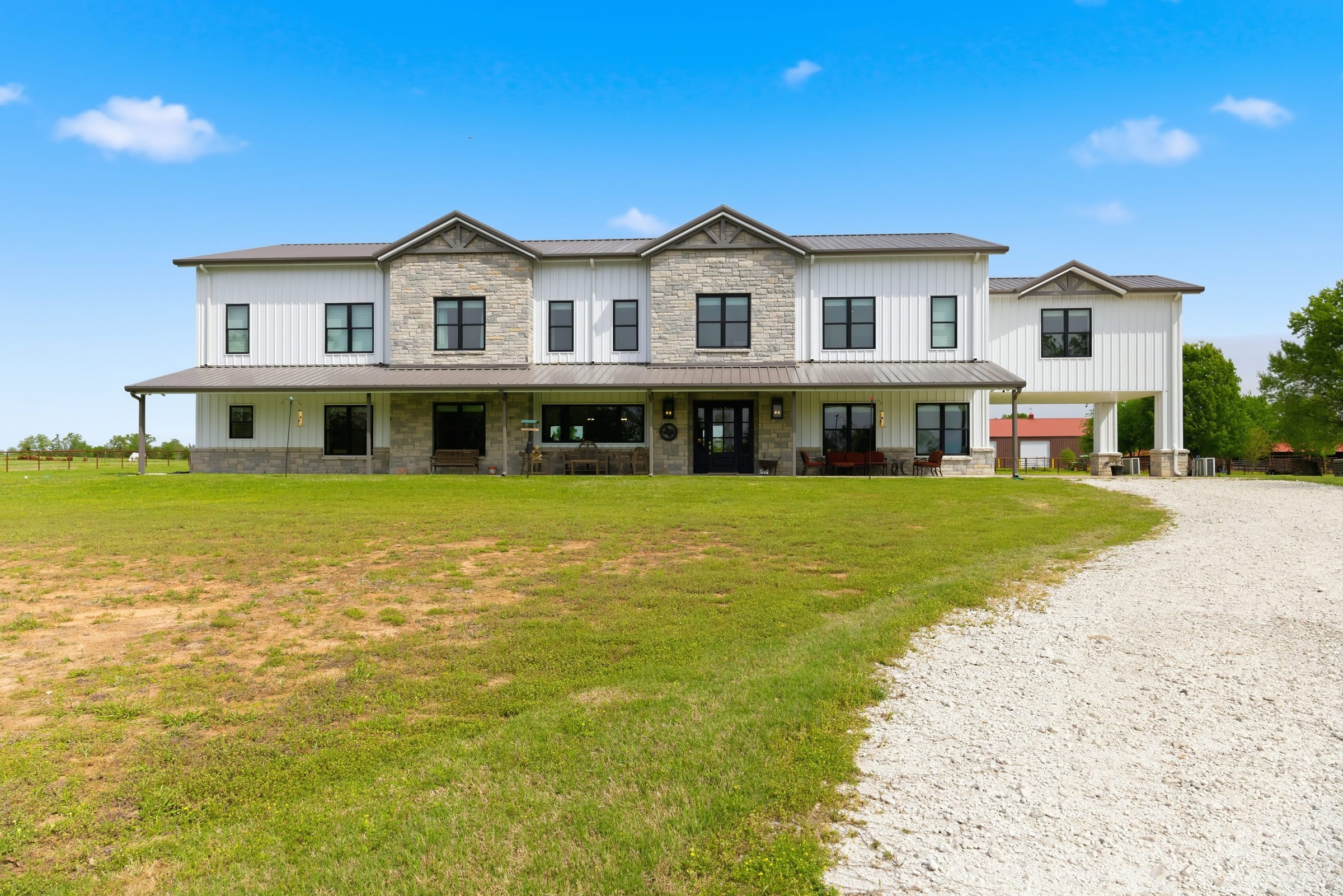 A two-storey white and stone ranch house seen across an open lawn, a gravel drive curving up to it under a blue sky.