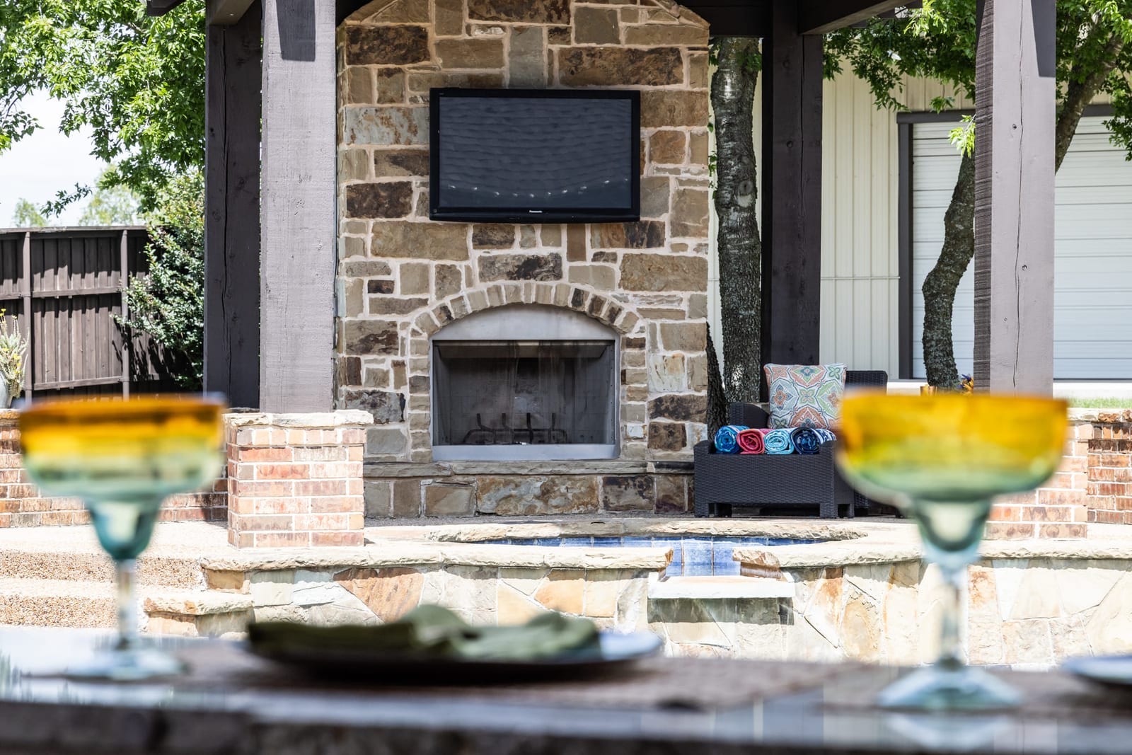 An outdoor stone fireplace and covered kitchen at dusk, two margarita glasses set on the bar in the foreground.