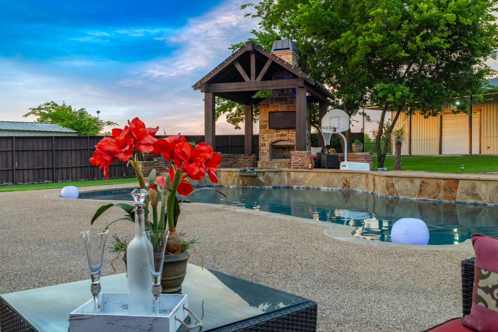 A backyard pool and stone cabana at dusk, a vase of red amaryllis on the glass table in the foreground.