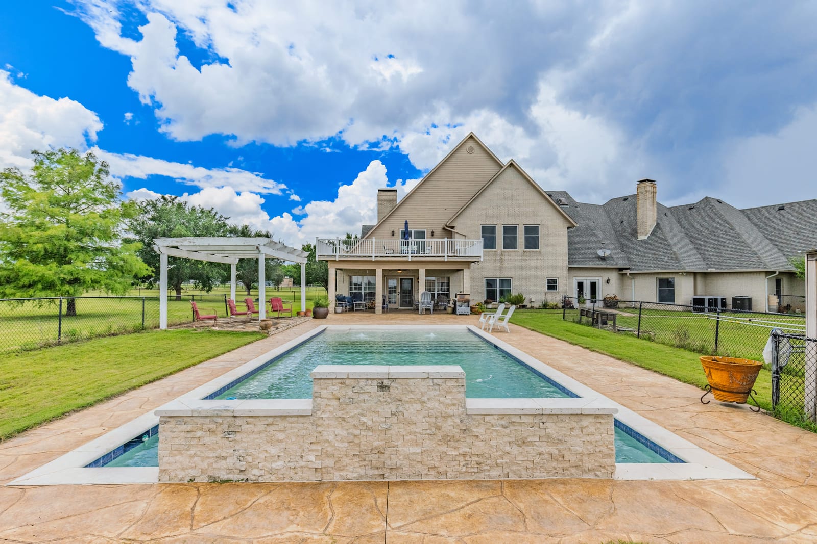 A rectangular pool with a stone water feature and a pergola, the house rising behind it under a cloud-marked sky.