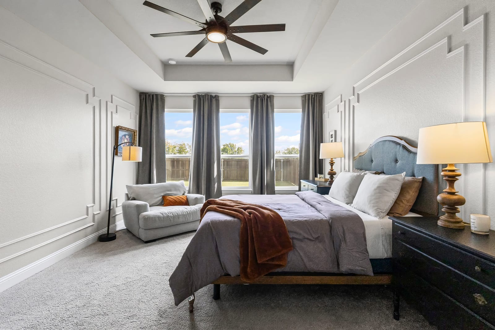 A staged primary bedroom in soft greys, a bench at the foot of the bed and daylight through a wall of windows.