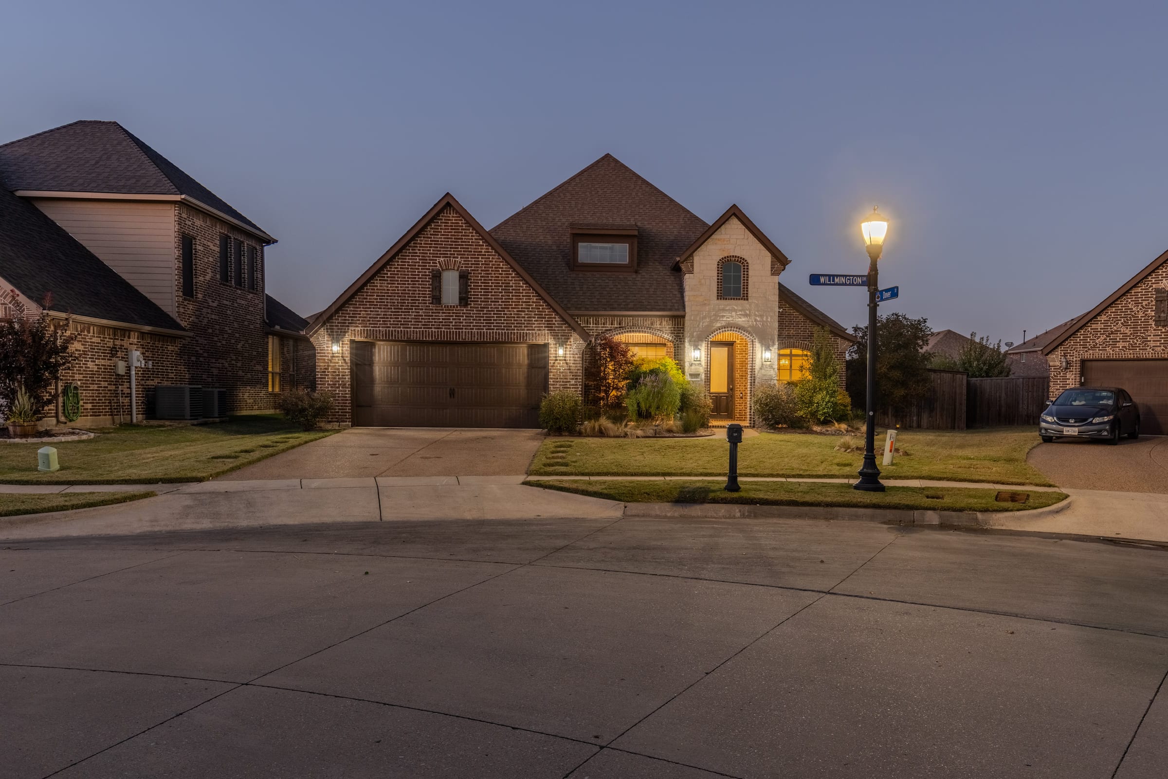 A two-storey brick house photographed from the street at dusk, windows lit and a streetlamp glowing at the kerb.