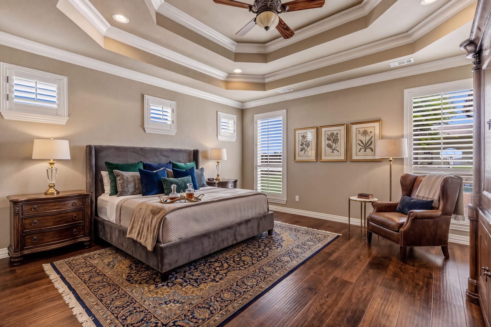 A primary bedroom under a coffered tray ceiling, hardwood floors and a leather armchair by the window.