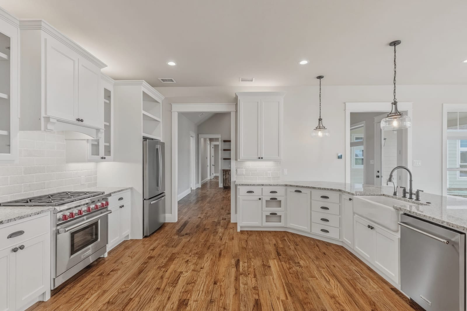 A white kitchen with a professional range, apron sink and pendant lights over warm wood floors.