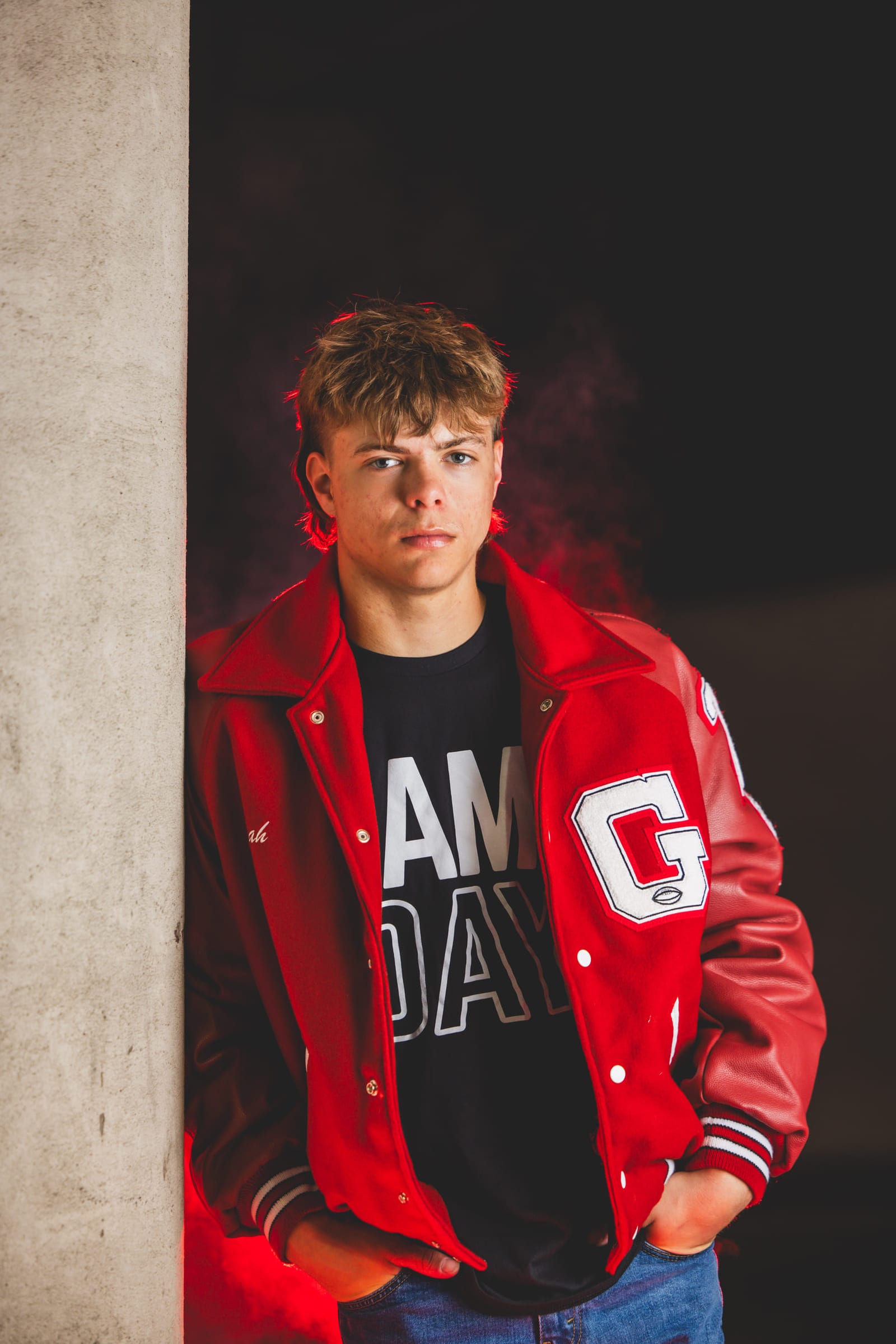 A senior in a red letterman jacket standing against a pale column, hands in his pockets.