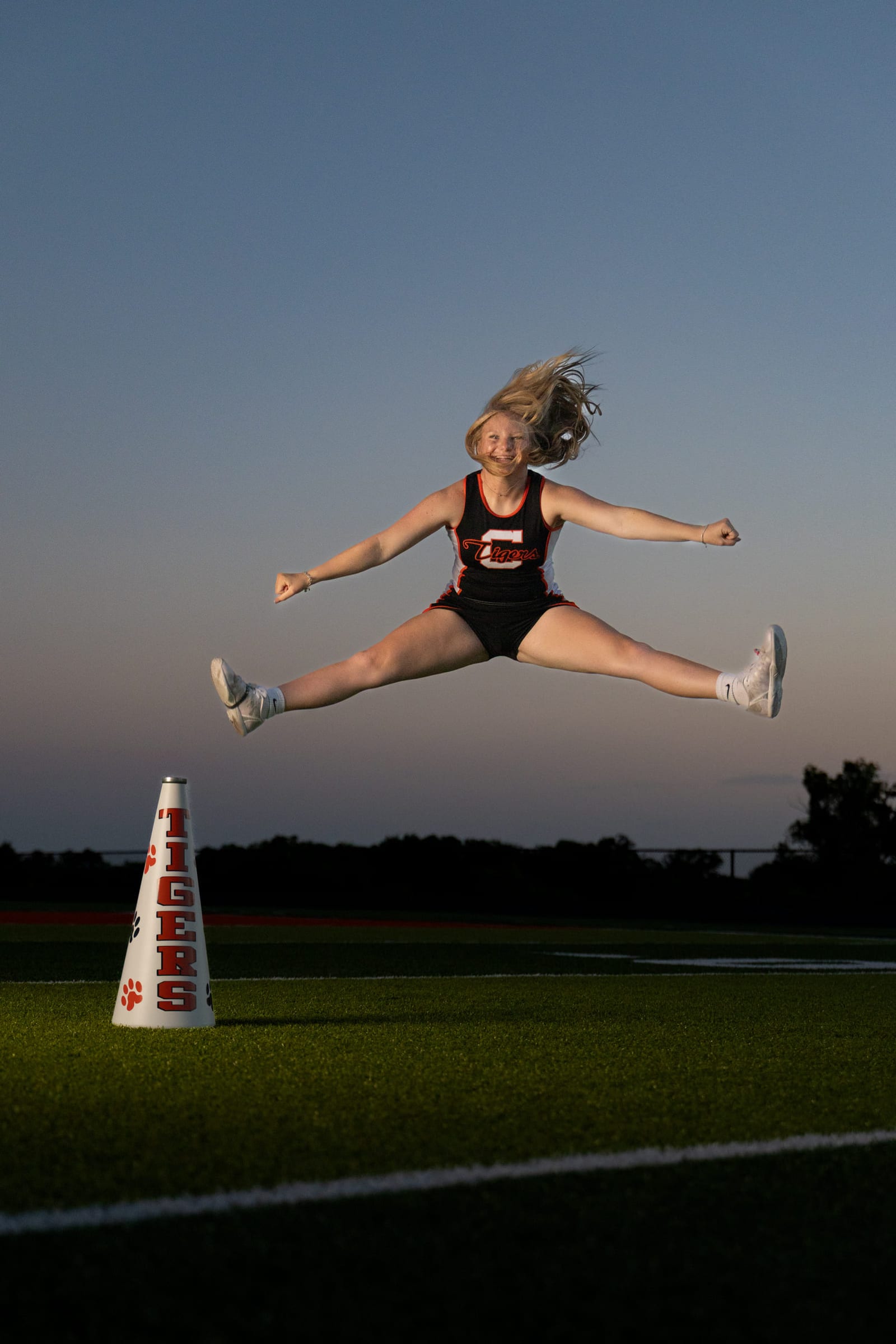 A cheerleader caught at the top of a toe-touch above a megaphone on a football field at dusk.