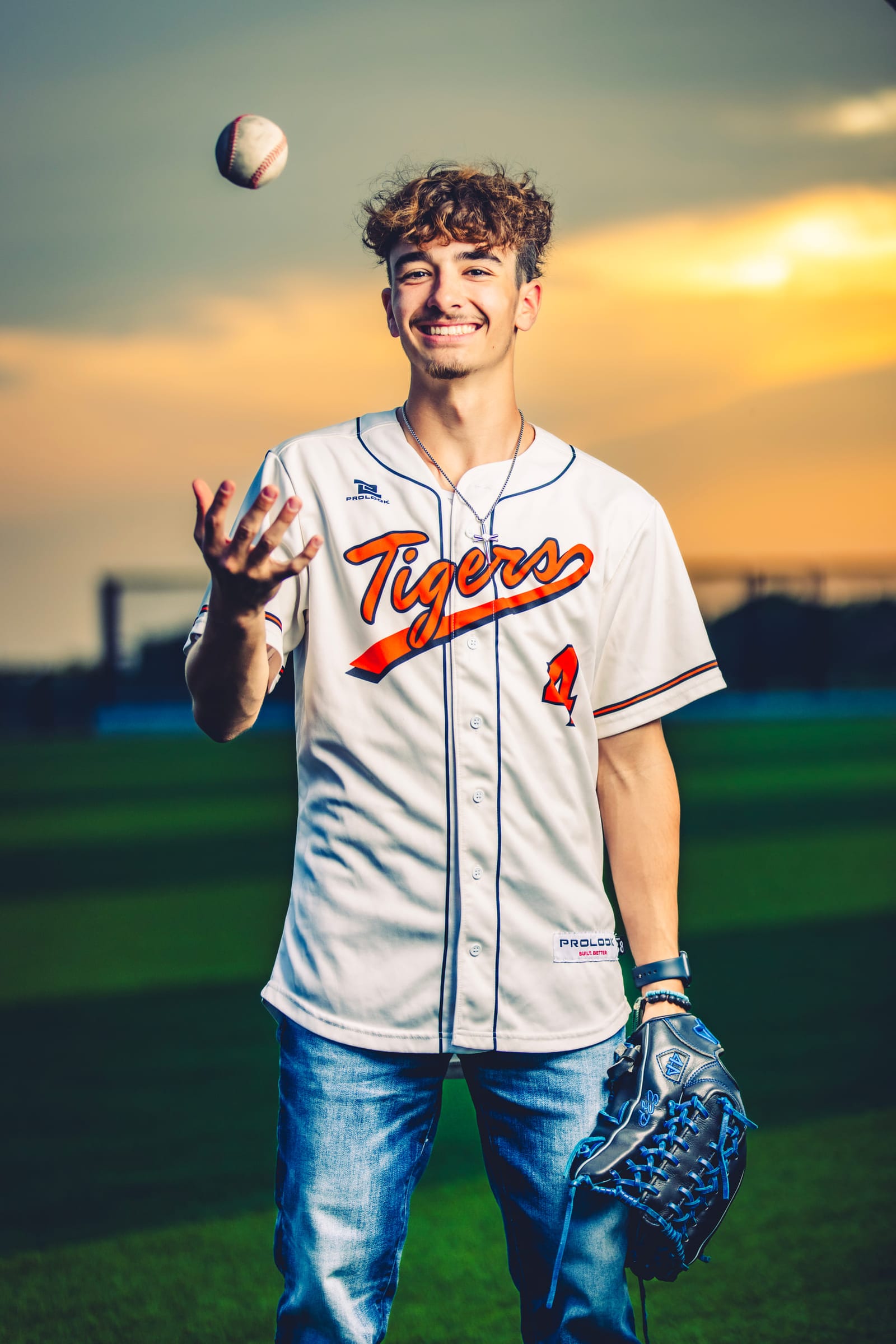 A senior in a baseball jersey tossing a ball above his head at sunset, glove in his other hand.