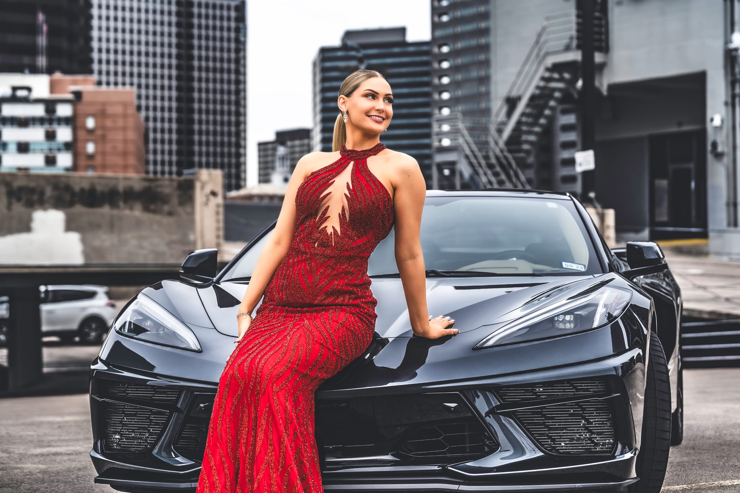 A senior in a red sequin gown seated on the hood of a Corvette, the city skyline behind.