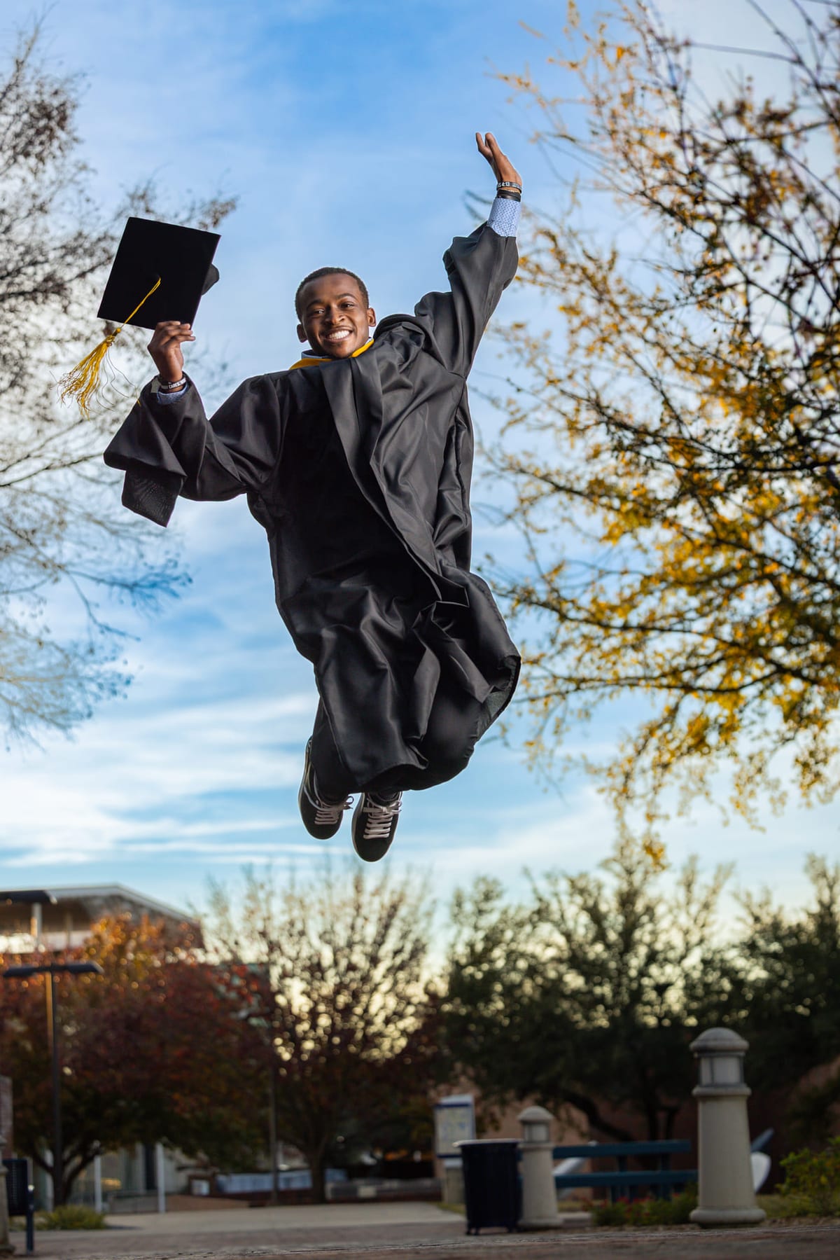 A graduate leaping off the ground in cap and gown, cap held high against a blue sky.