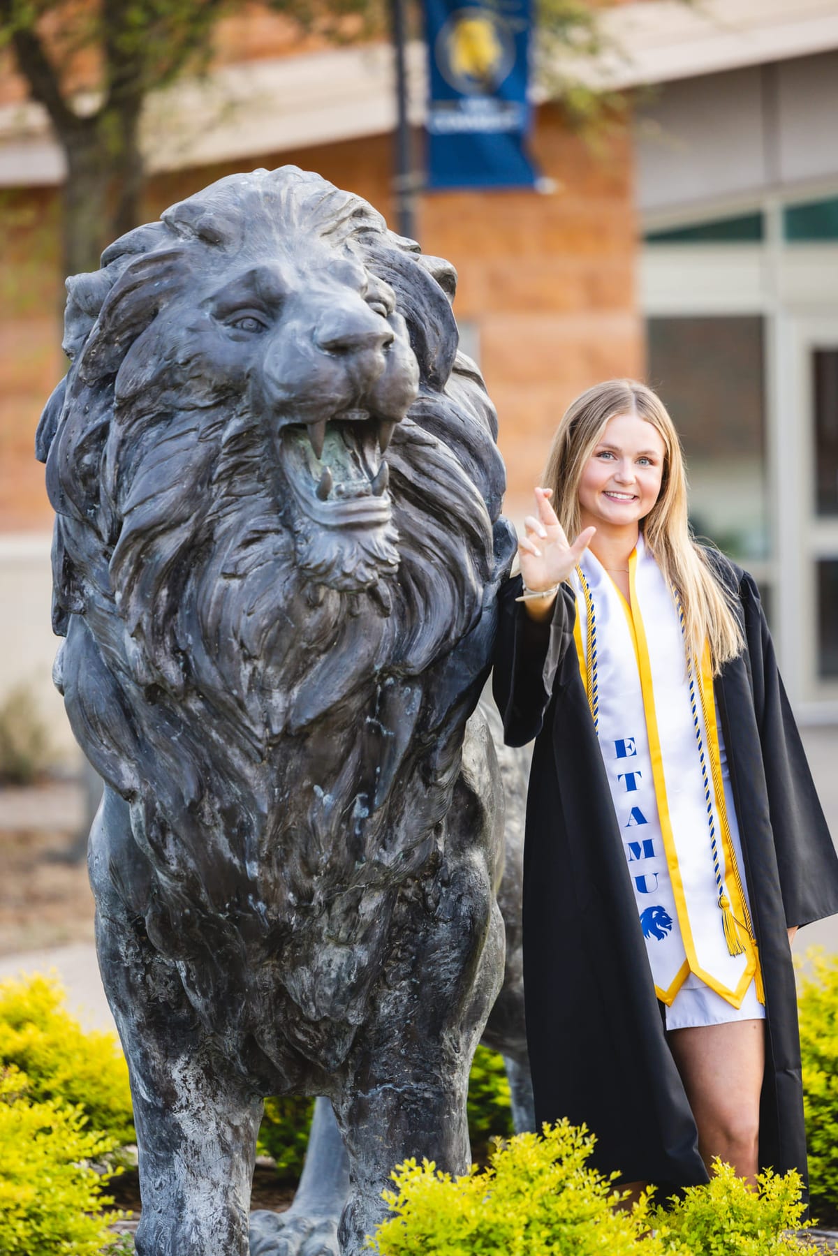 A graduate in cap, gown and honour cords beside a bronze lion statue on campus.