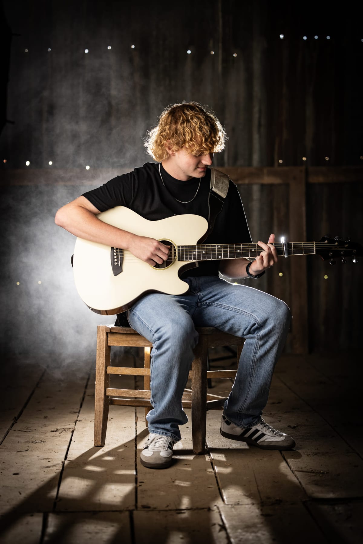 A senior playing an electric guitar on a stool in a barn, haze lit behind him.