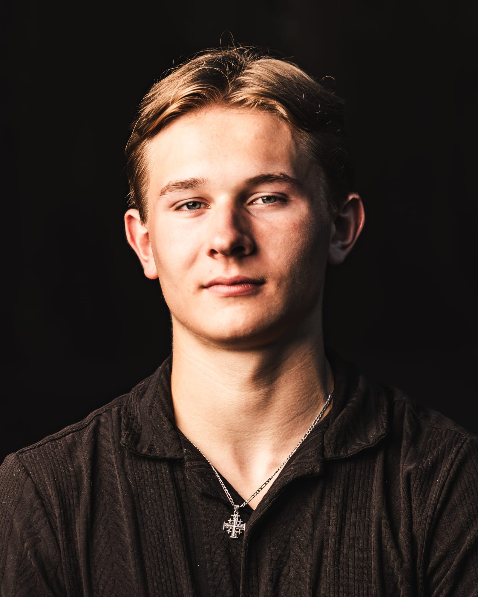 Studio portrait of a senior in a dark shirt on a black ground, a single light raking across his face.