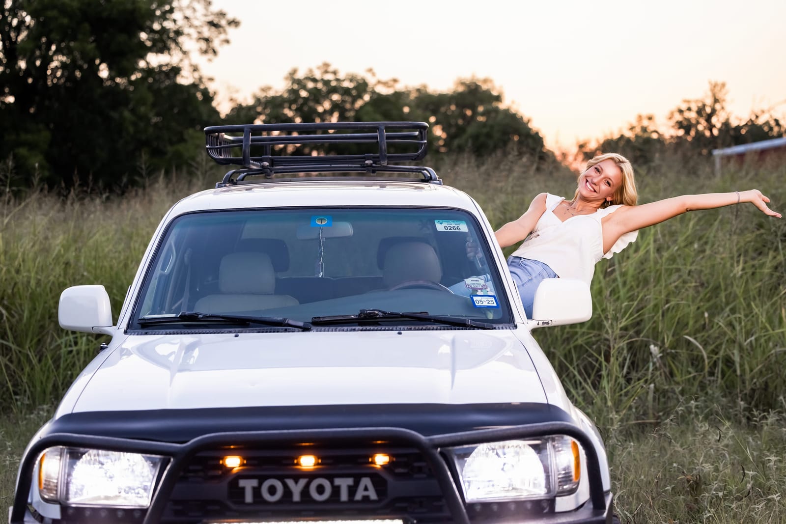 A senior leaning out of a Land Cruiser’s window with both arms open, headlights on at sunset.