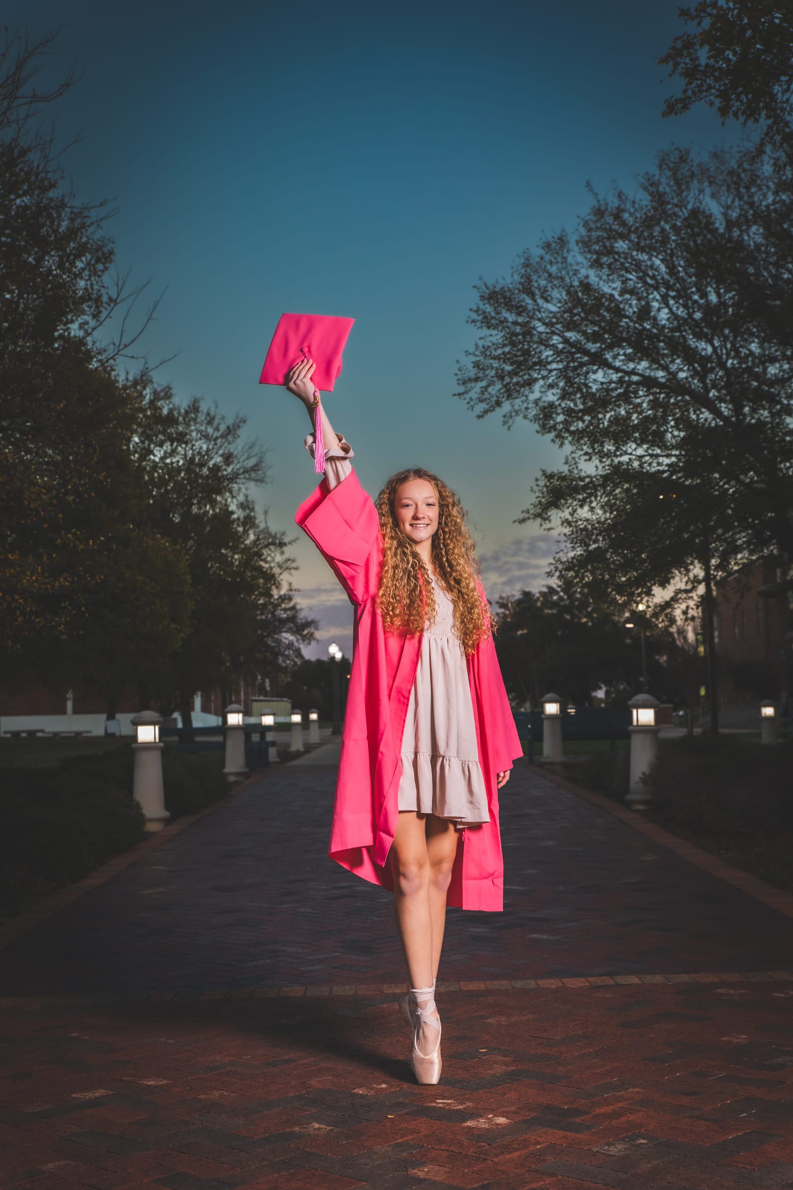 A graduate in a pink gown walking a brick avenue at blue hour, cap held up in one hand.