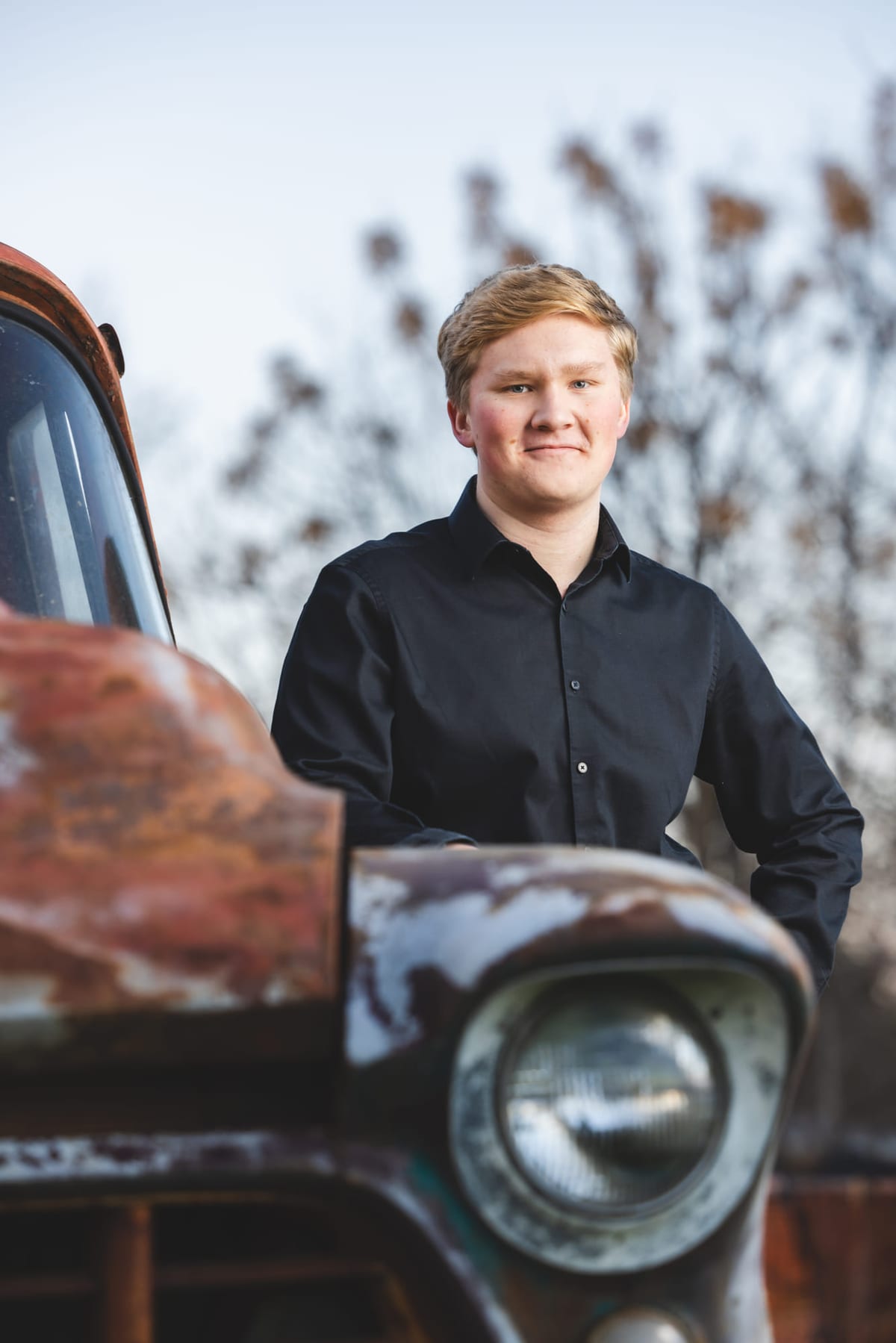 A senior in black leaning against the wing of a rusted pickup, chrome headlamp in the foreground.