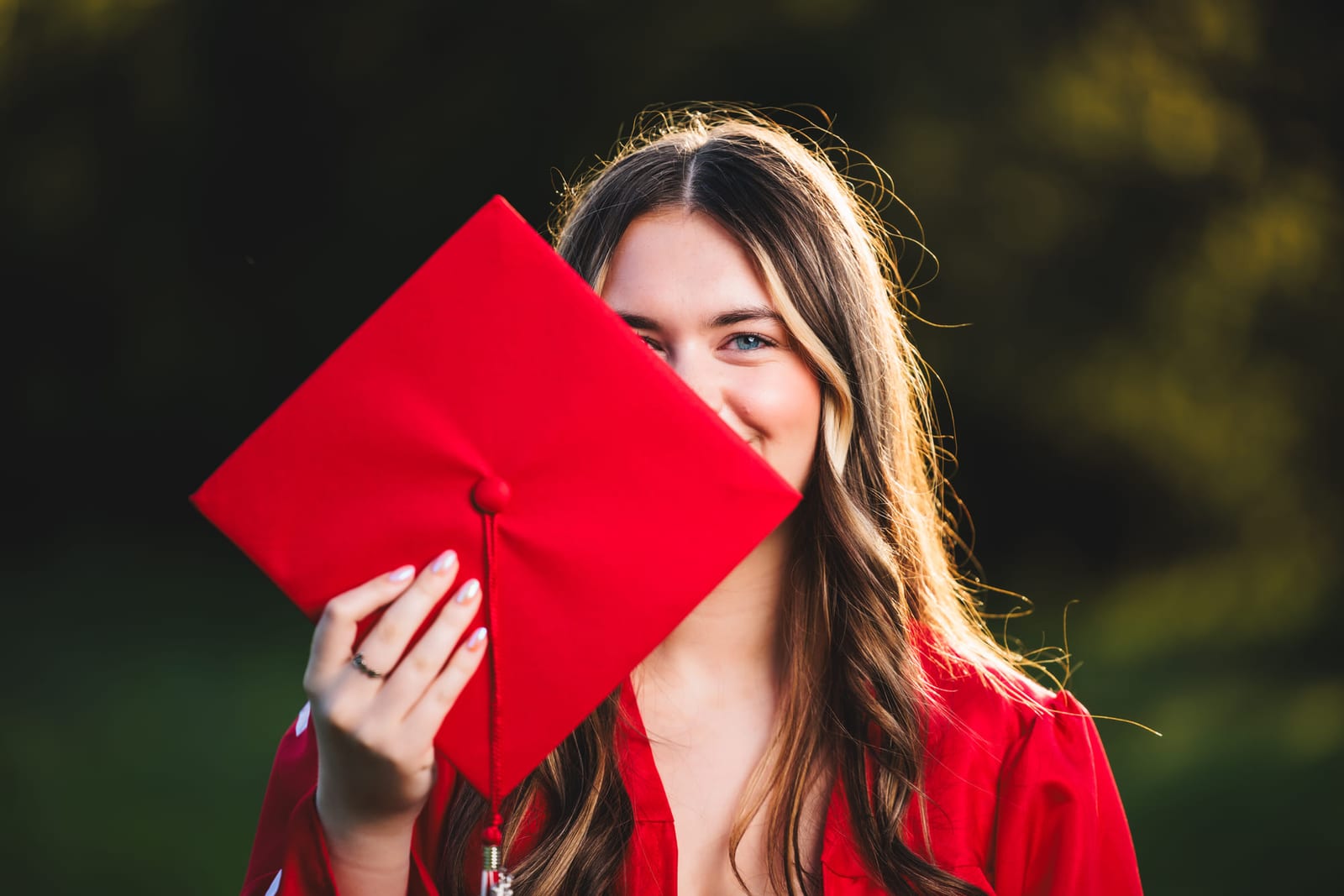 A graduate holding a red mortarboard in front of her face, one eye showing above the brim.