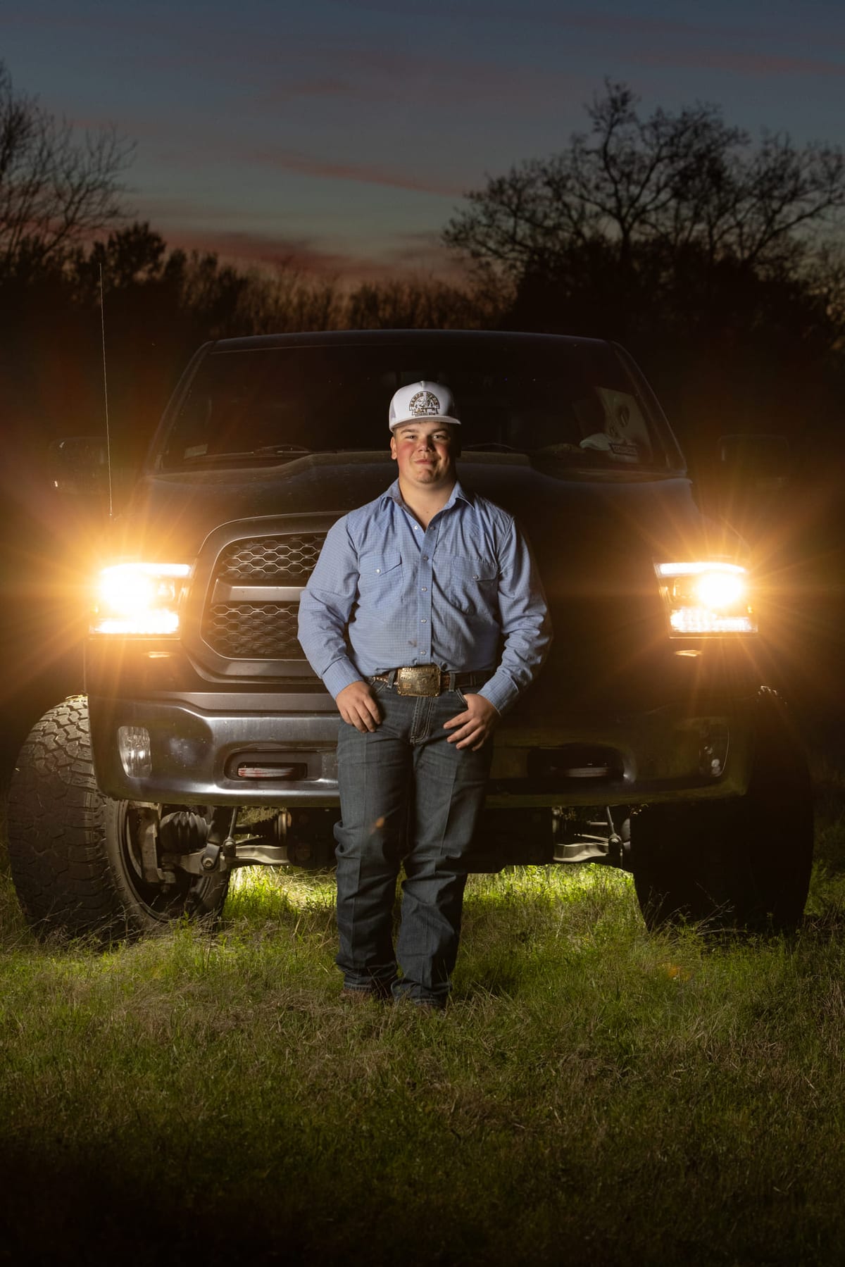 A senior standing in front of a lifted pickup at dusk, both headlights burning.
