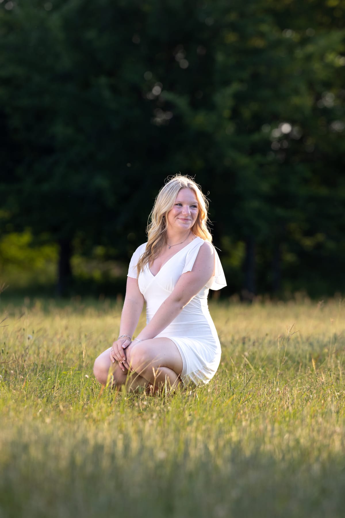A senior kneeling in a sunlit meadow in a white dress.