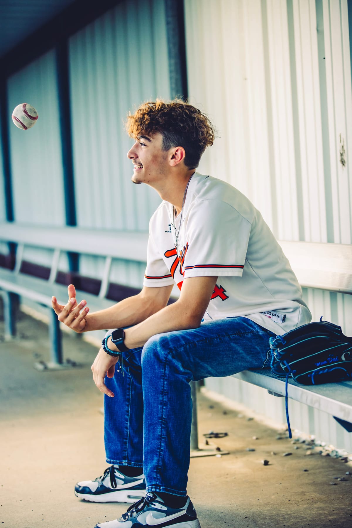 A ballplayer sitting on a dugout bench juggling two baseballs.
