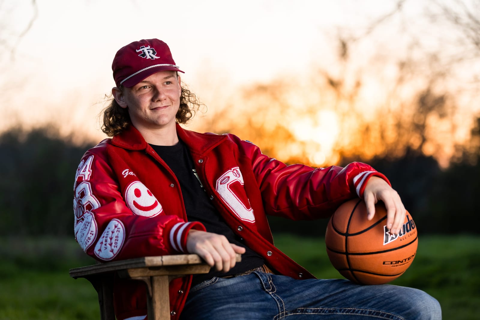 A player in a letterman jacket seated with a basketball at sunset.