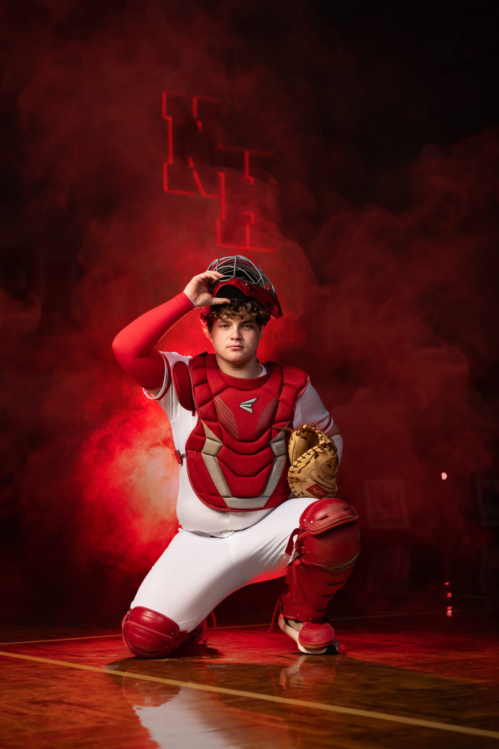 A catcher kneeling in full gear, red smoke lit behind him on the gym floor.
