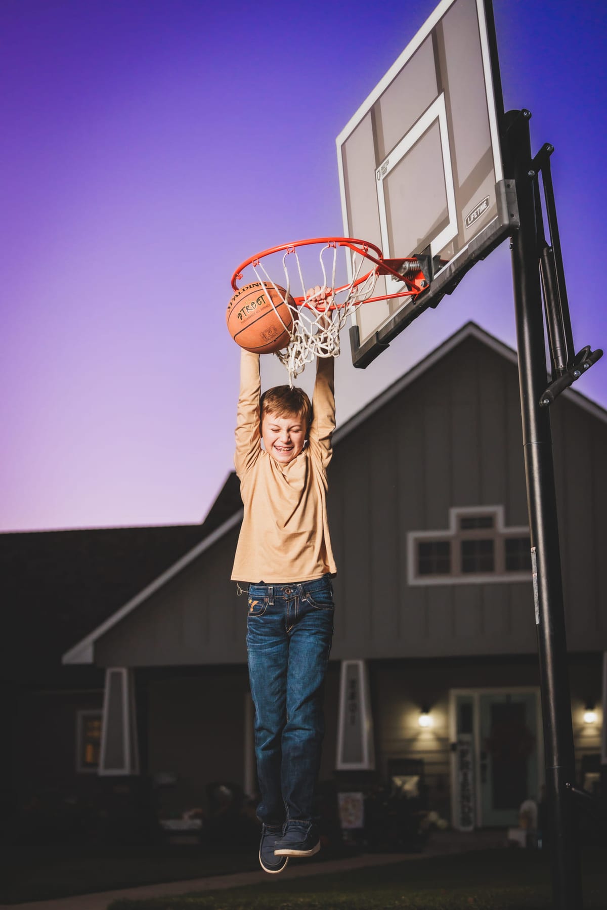 A young player hanging from the rim of a barn-side hoop against a violet sky.