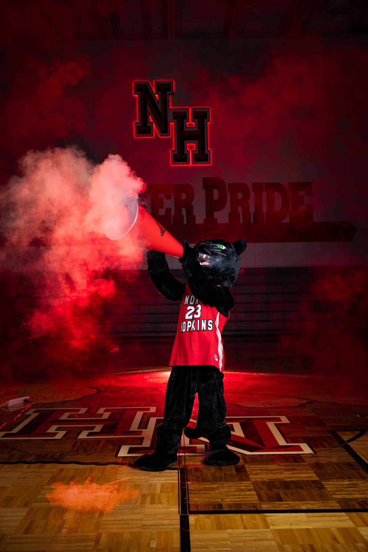 A school mascot on the gym floor releasing red smoke, team lettering on the wall behind.