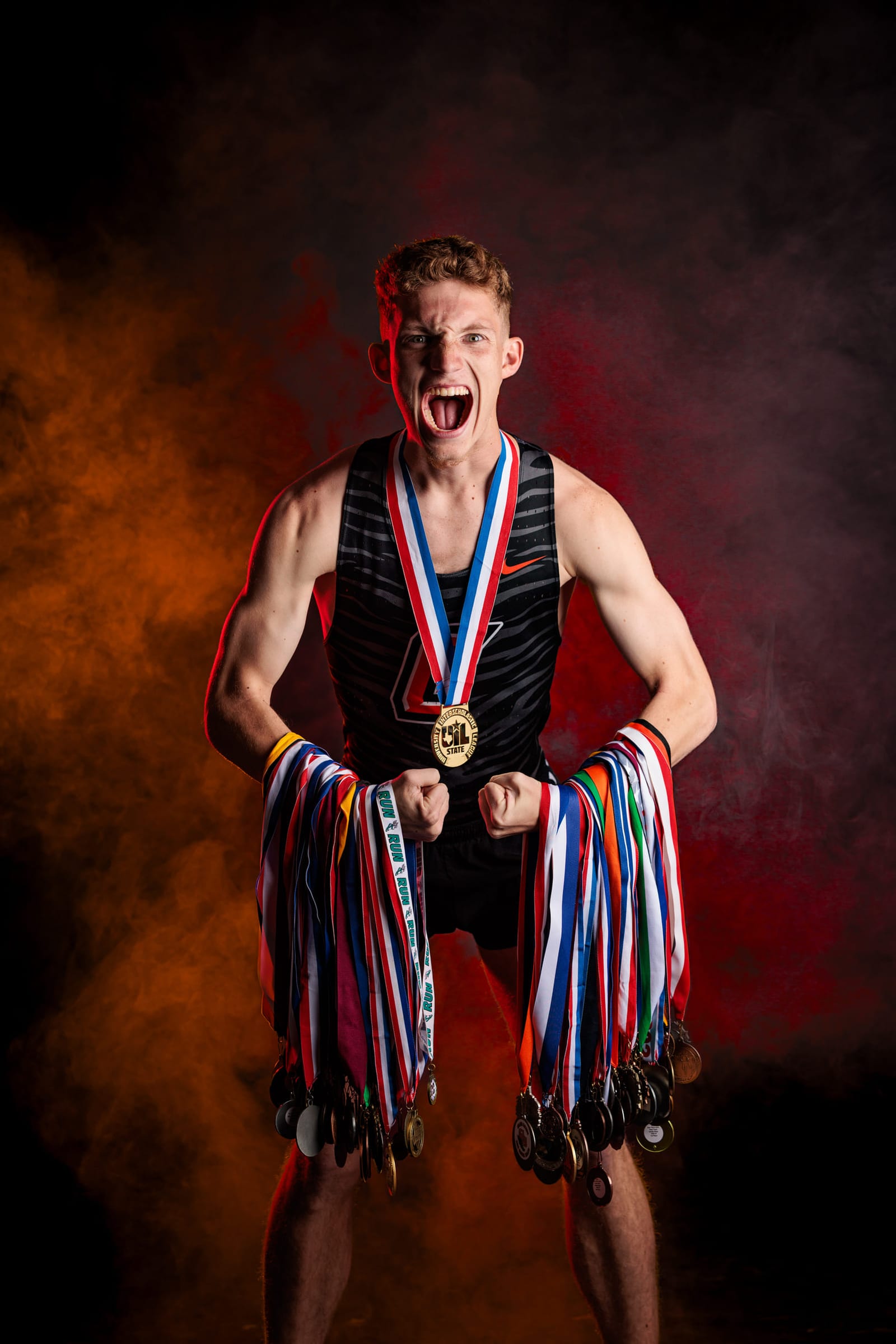 An athlete in a singlet holding out fistfuls of medals, orange smoke behind.
