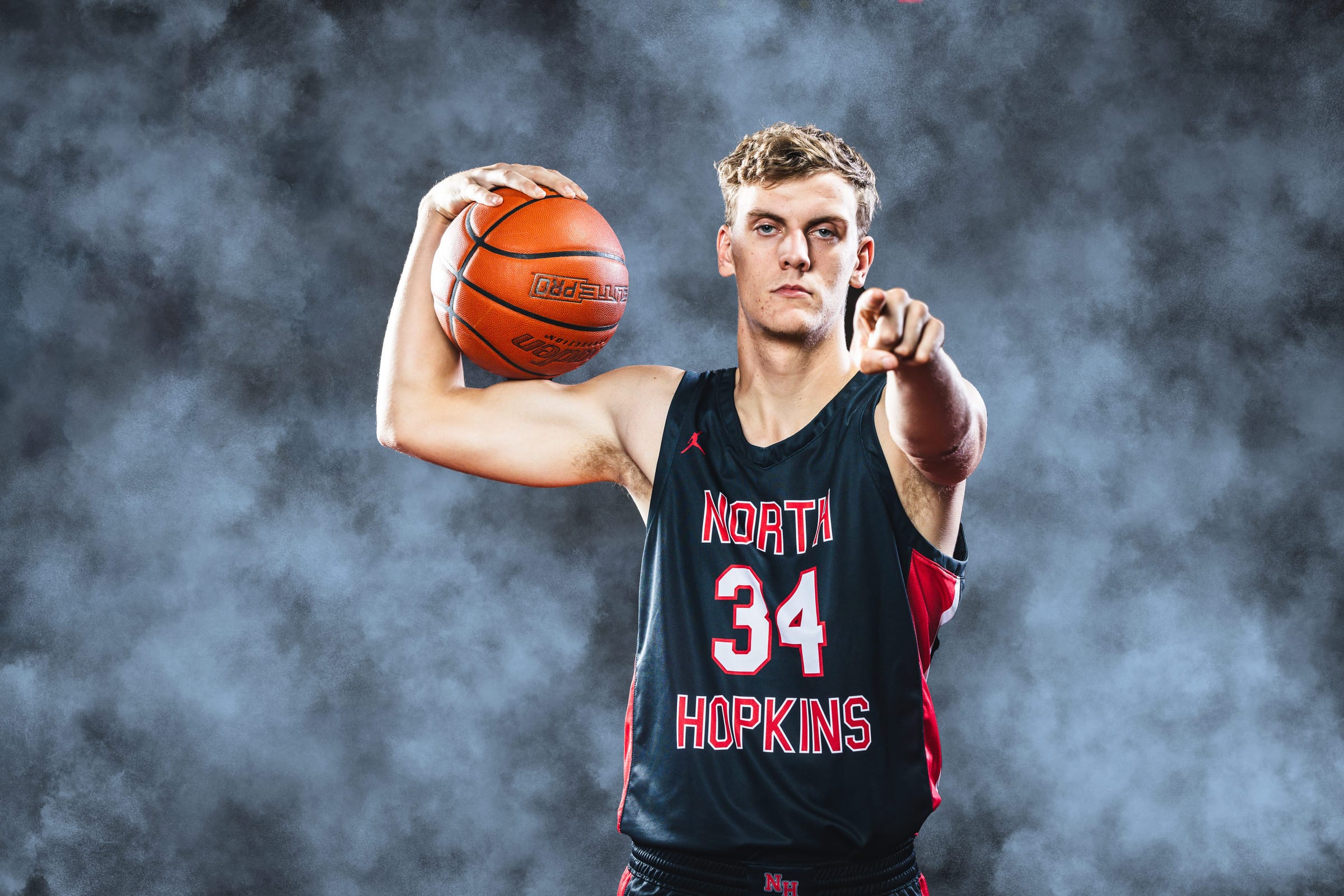 A high-school basketball player holding the ball at his shoulder, pointing at the camera through grey studio haze.