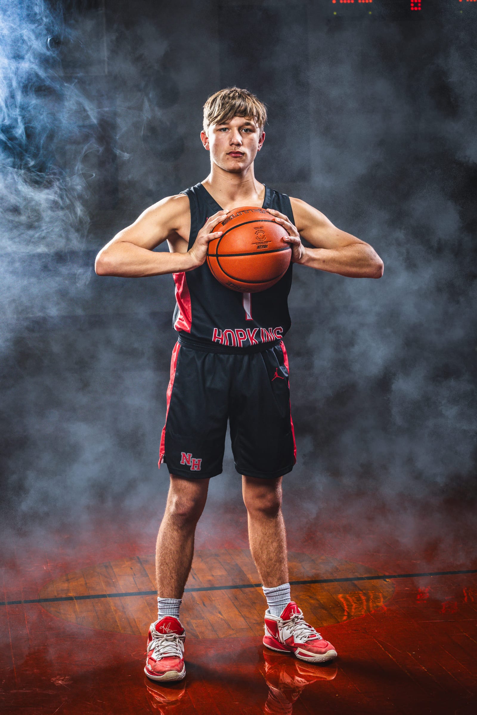 A basketball player standing full length on a court, ball held at his chest, smoke rolling behind him.