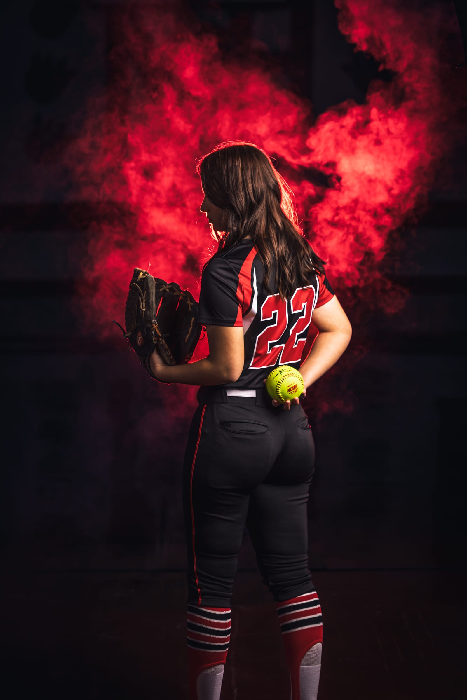 A softball player photographed from behind, glove and ball down at her sides, red smoke lit around her.