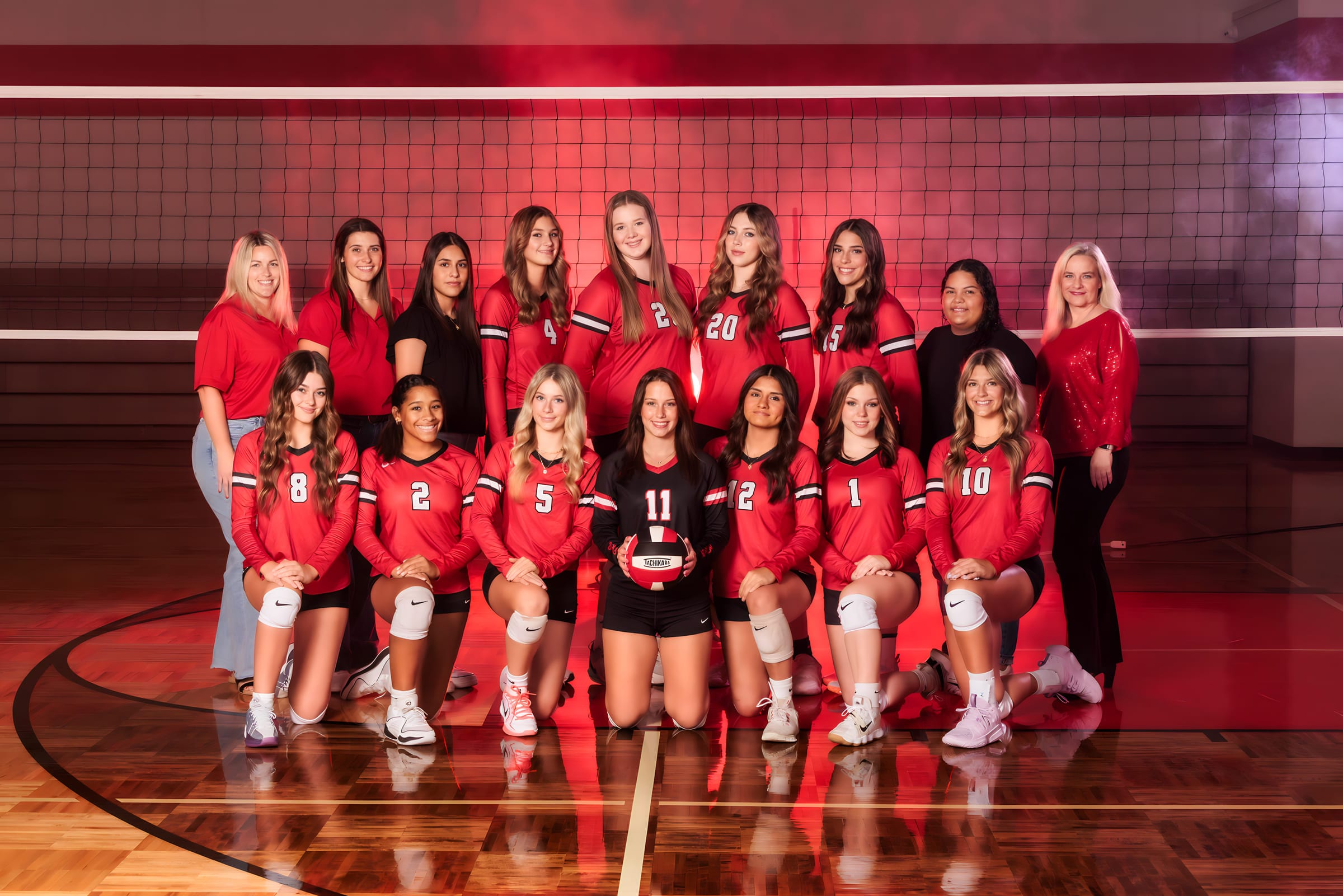 A varsity volleyball squad posed across the gym floor under red light.